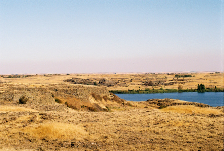 [Mostly flat land covered in very short tan grasses beside a body of water.]